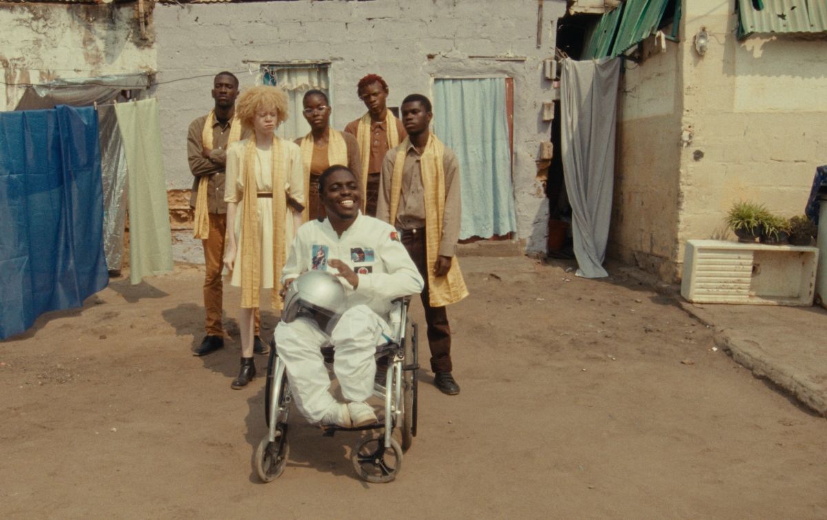 Isis Hembe as Man Ré in The Adventures of Angosat, seated in a wheelchair with performers in Cazenga, Luanda.