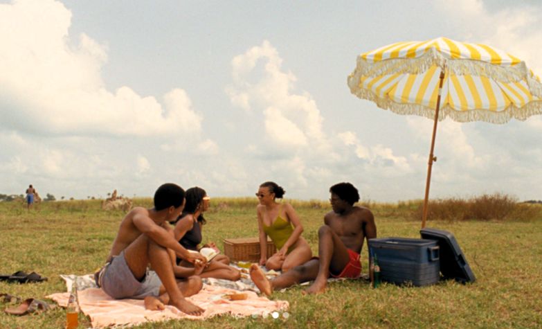A still from "Clarissa" (2026) — a group of young friends on a picnic blanket under a yellow-and-white striped umbrella in a sunlit Lagos field